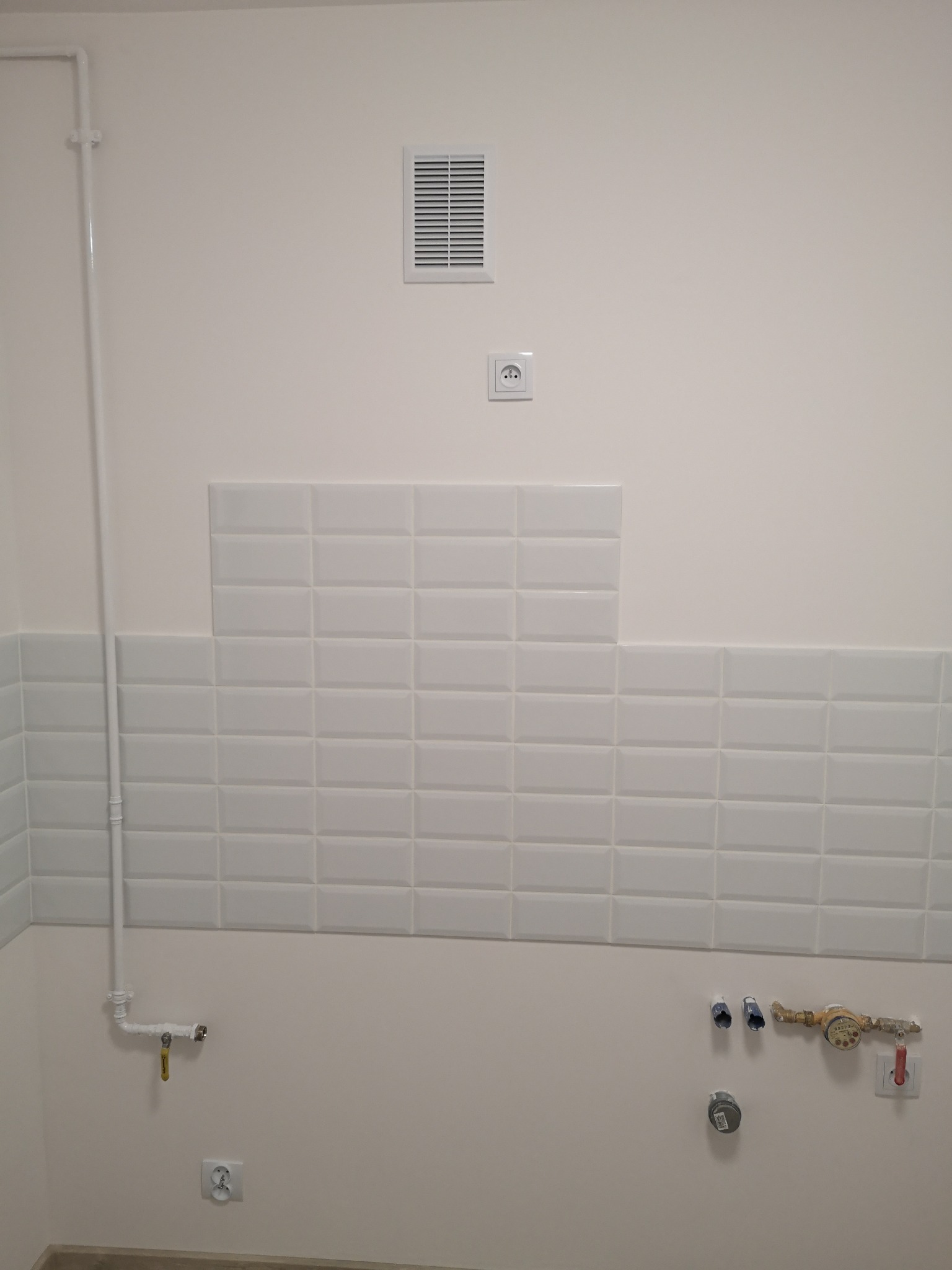 Fragment ściany z częściowo ułożonymi białymi, prostokątnymi płytkami ceramicznymi, widoczne instalacje hydrauliczne, wentylacja i gniazdko elektryczne.