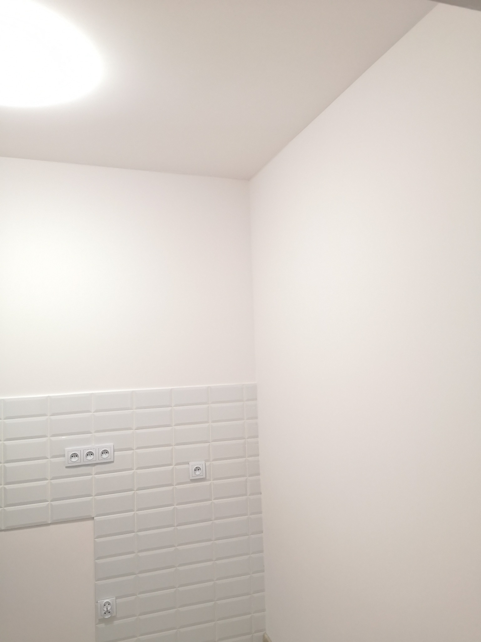 Świeżo wykończona, biała ściana z białymi, prostokątnymi płytkami, na których zamontowano gniazdka elektryczne w białych ramkach, z fragmentem ściany bez płytek, oświetlona centralnym światłem.