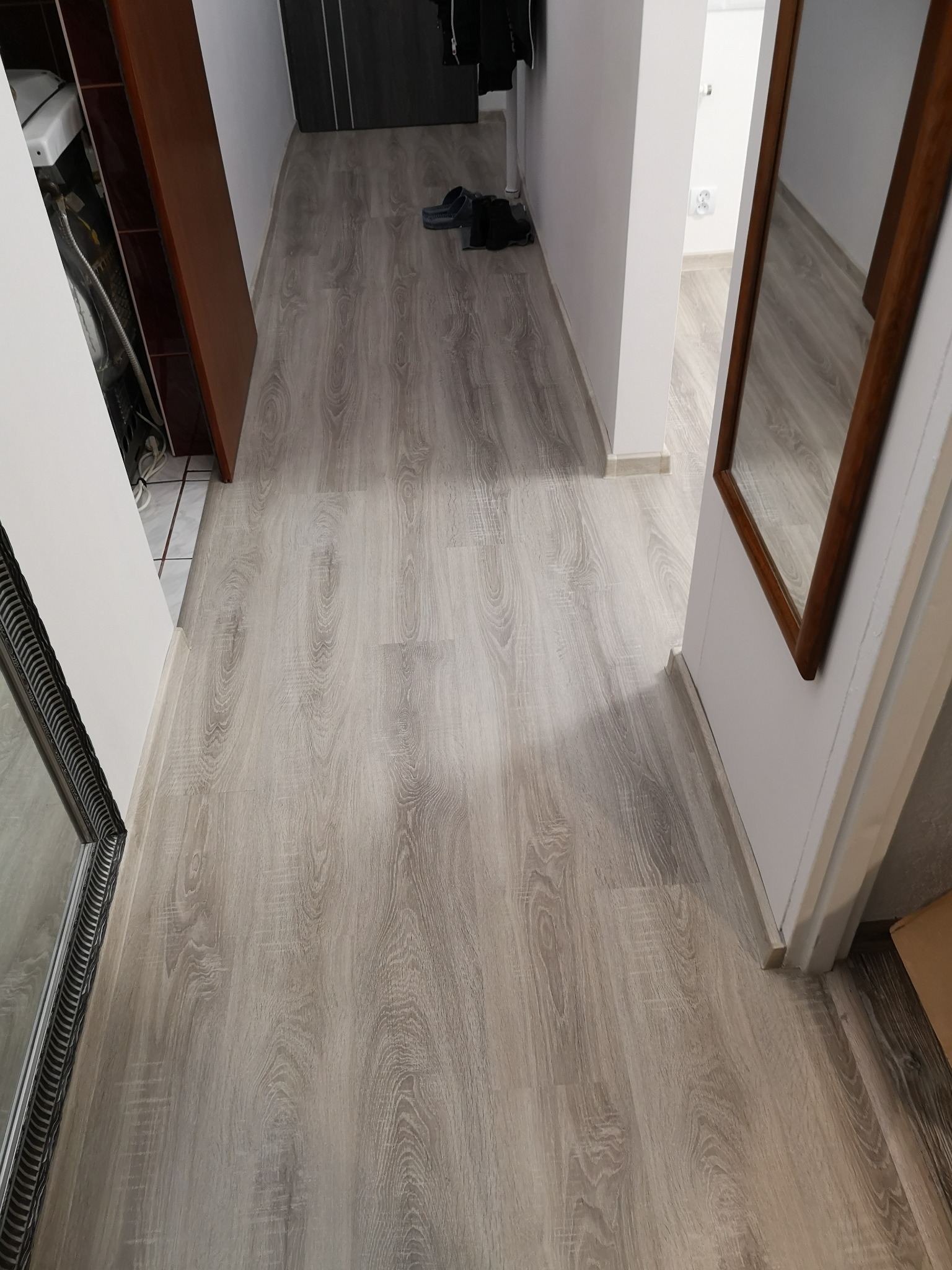 Jasne panele podłogowe imitujące drewno w korytarzu, widoczne buty i fragment lustra w ozdobnej ramie.