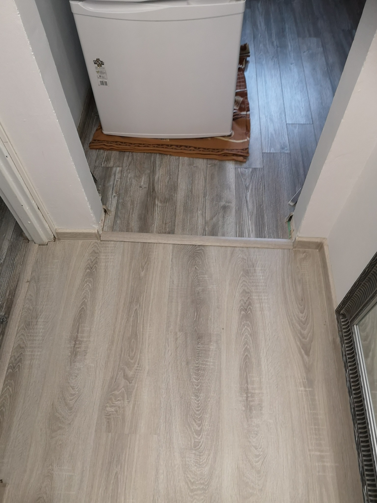 Fragment podłogi wyłożonej panelami w odcieniach szarości, widoczny próg drzwiowy i fragment ściany z niewielką lodówką w tle.