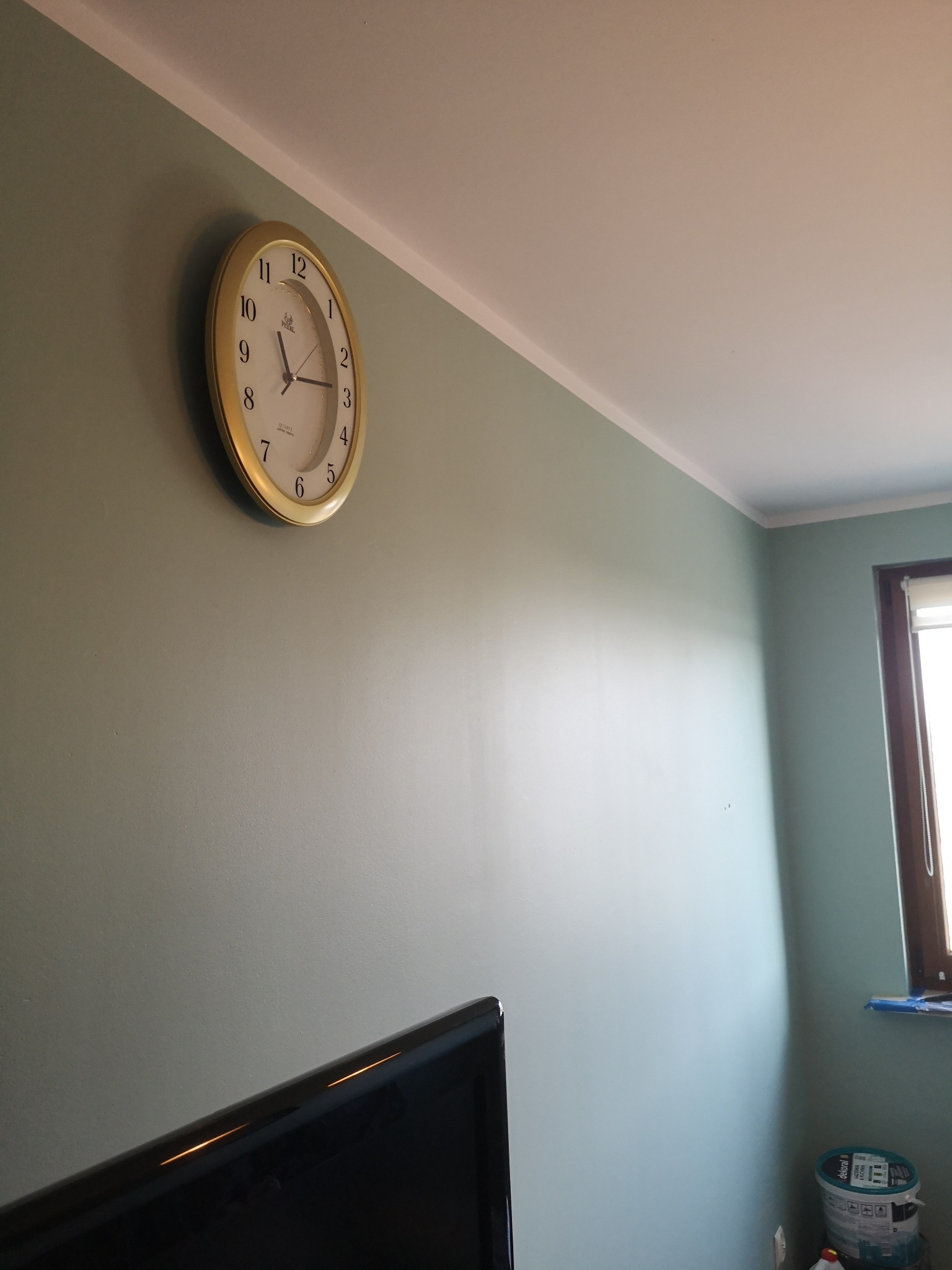 Świeżo pomalowane ściany w odcieniach zieleni z zamontowanym zegarem ściennym, widoczny fragment telewizora i puszka farby w rogu pokoju.