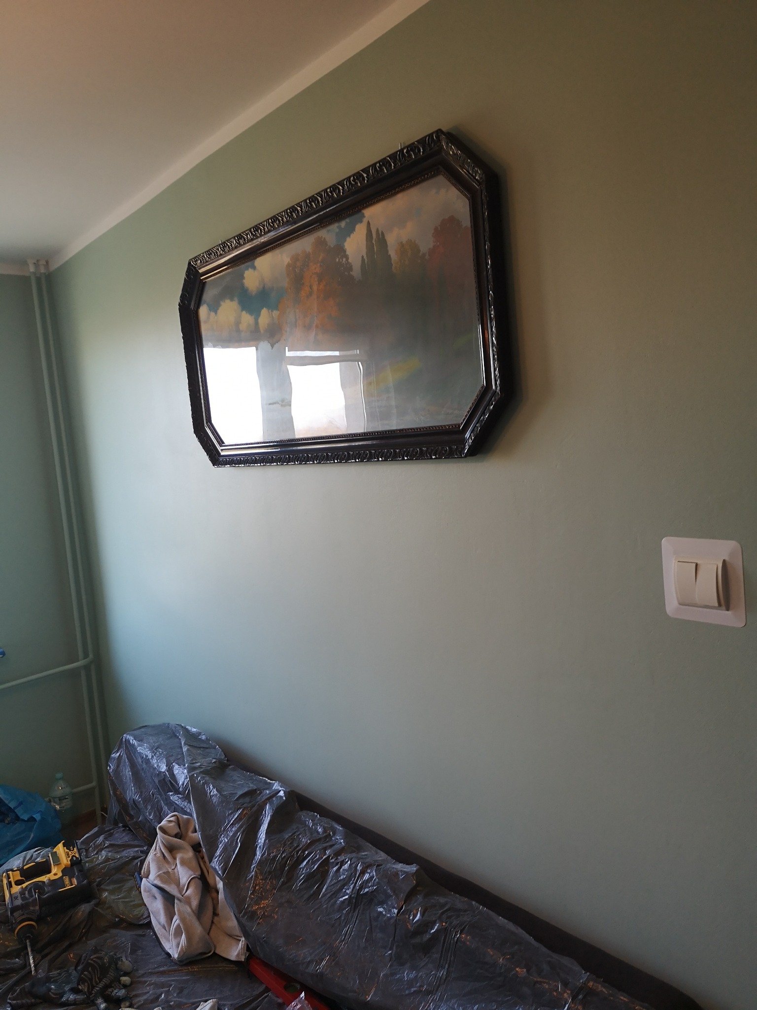 Pokój w trakcie przygotowań do malowania: ściana w odcieniach zieleni, duży obraz w ozdobnej ramie, zabezpieczone folią meble, widoczna wiertarka i akcesoria malarskie.