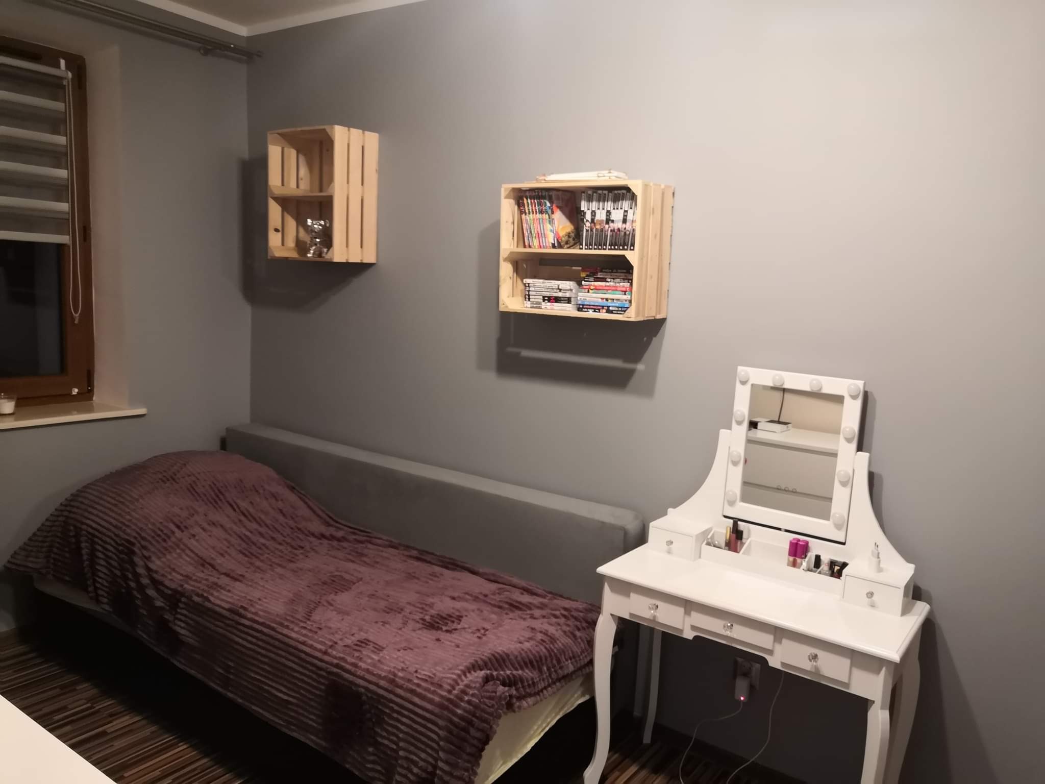 Pokój z szarą ścianą, drewnianymi półkami z książkami i ozdobami, łóżkiem z bordową narzutą oraz białą toaletką z lustrem podświetlanym żarówkami.
