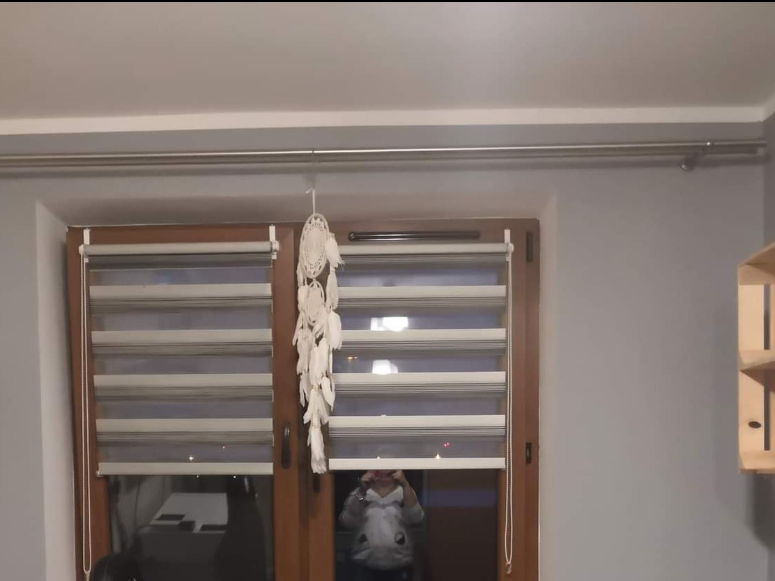 Okno z drewnianą ramą i roletami dzień-noc, ozdobione łapaczem snów z białych piór.