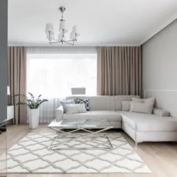Jasny salon z narożną sofą, szklanym stolikiem kawowym i wzorzystym dywanem, oświetlony nowoczesnym żyrandolem.