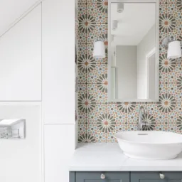 Nowoczesna łazienka z białą, okrągłą umywalką nablatową, baterią chromowaną, lustrem w ramie i ścianą wyłożoną dekoracyjnymi, marokańskimi płytkami w odcieniach beżu, brązu i błękitu. Po lewej...