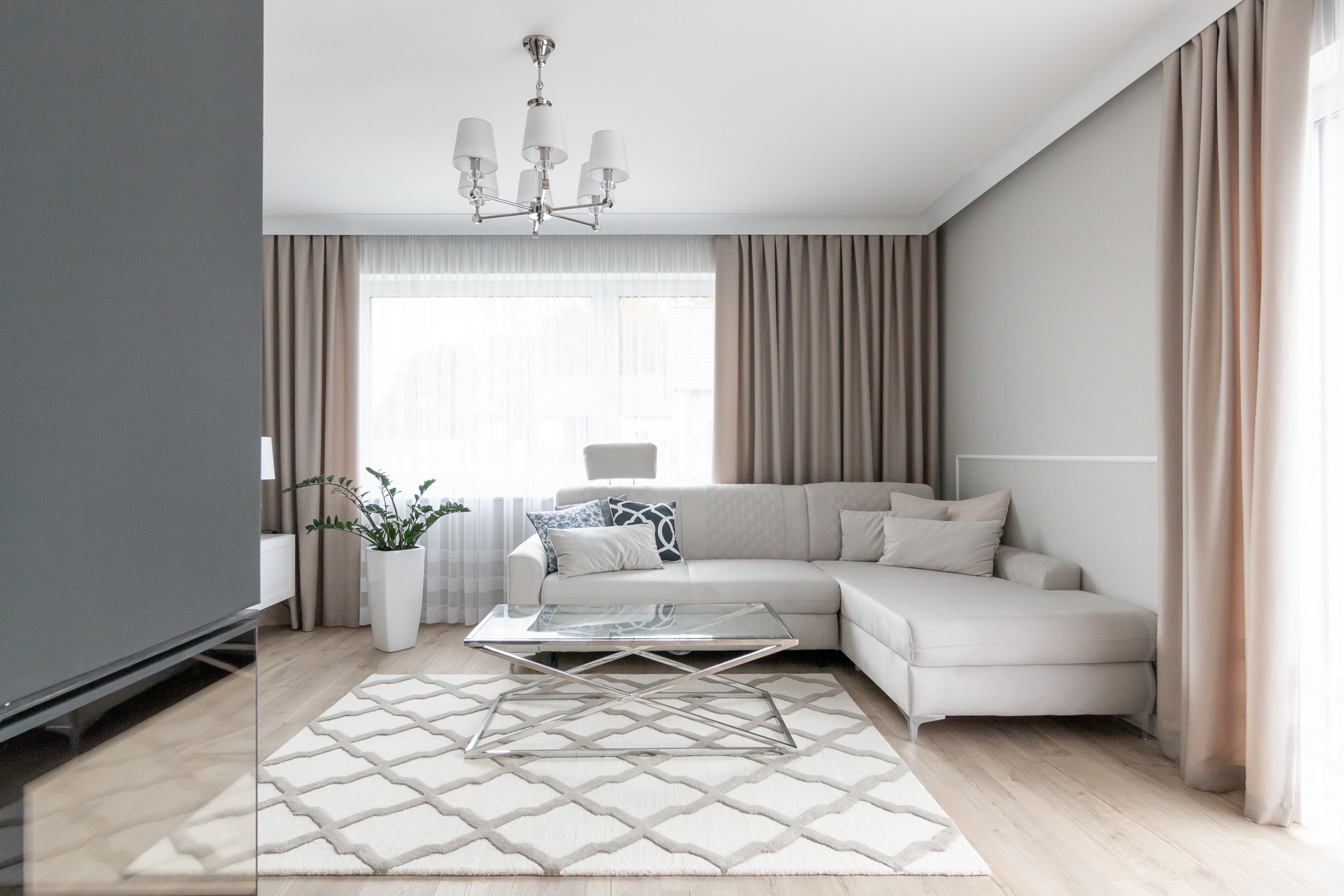Jasny salon z narożną sofą, szklanym stolikiem kawowym na metalowym stelażu, wzorzystym dywanem i rośliną w białej donicy, oświetlony przez okno z beżowymi zasłonami i nowoczesny żyrandol.