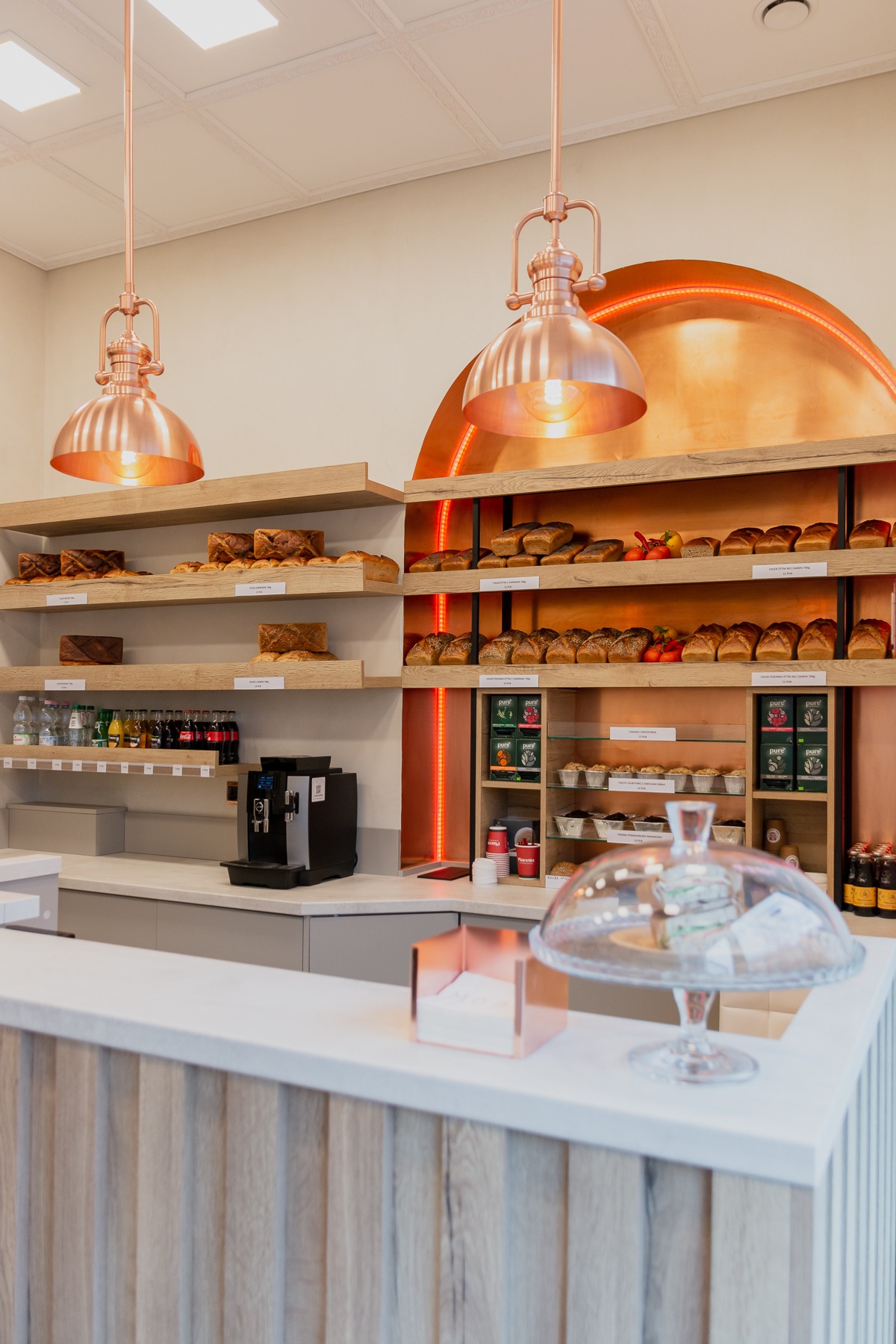 Wnętrze lokalu usługowego: jasna lada z drewnianym frontem, za nią półki z pieczywem, napojami i kawą, podświetlane miedzianą ścianą w kształcie łuku, nad ladą wiszą miedziane lampy.