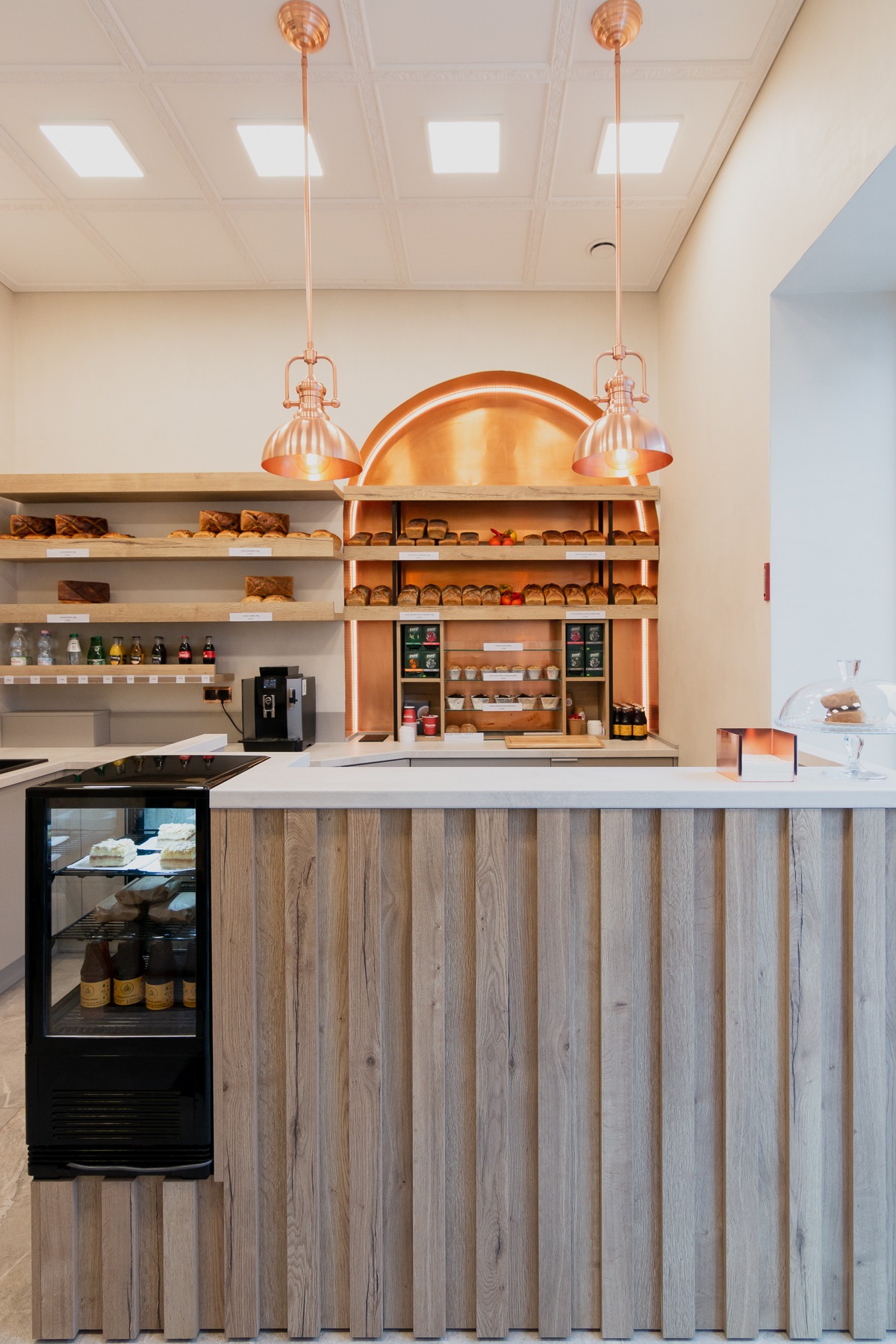 Wnętrze piekarni lub kawiarni z drewnianą ladą, miedzianymi lampami i półkami pełnymi pieczywa oraz napojów w chłodziarce.