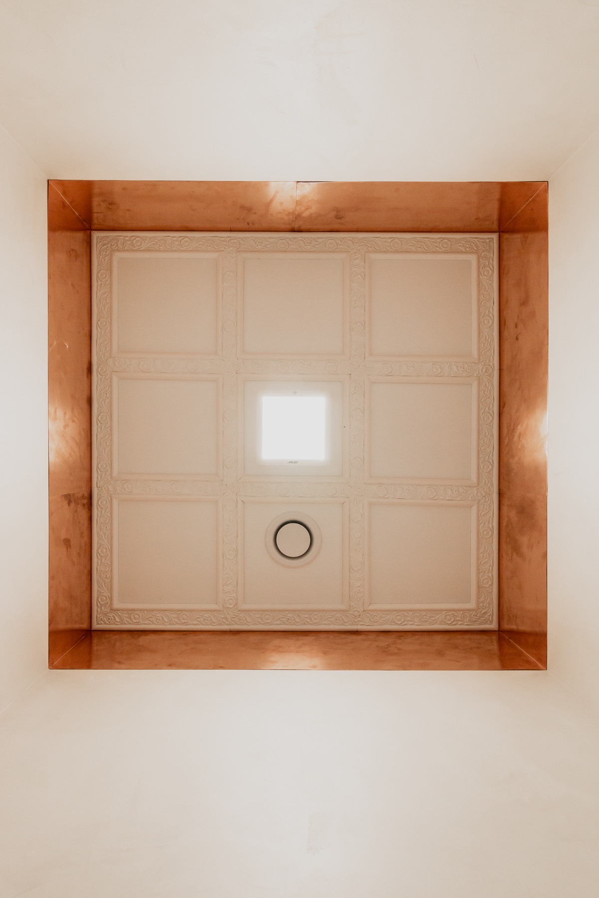 Kasetonowy sufit z dekoracyjnymi obramowaniami w stylu klasycznym, oświetlony centralnie kwadratową lampą i otoczony miedzianą listwą.