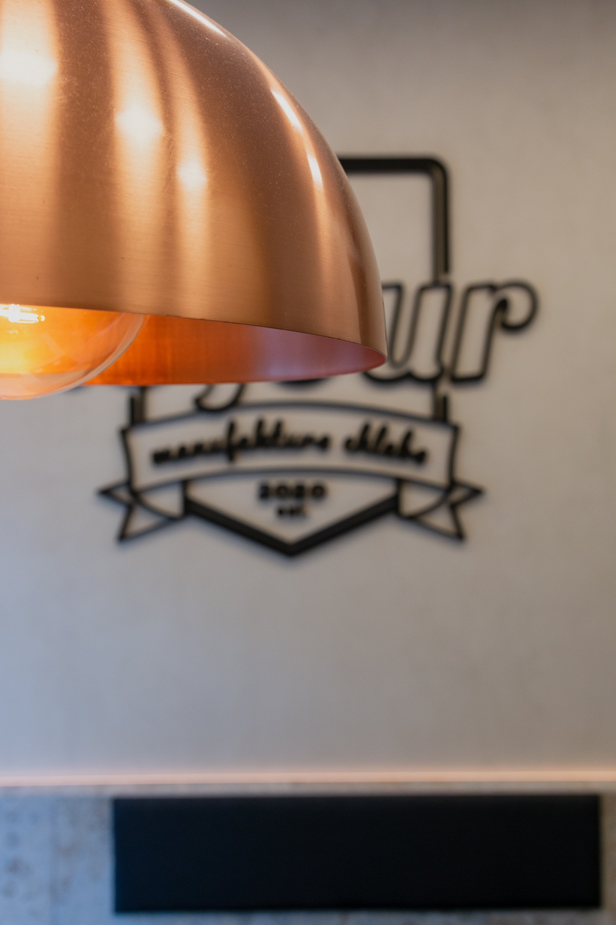 Miedziana lampa wisząca nad ścianą z logo piekarni 'Your manufaktura chleba 2020'.