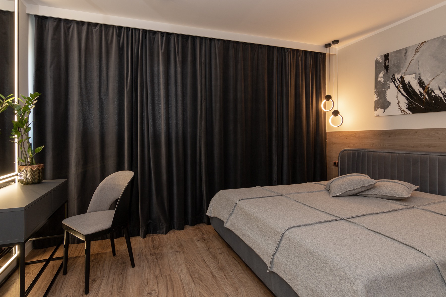 Nowoczesna sypialnia w odcieniach szarości z drewnianą podłogą, charakteryzująca się czarnymi, welurowymi zasłonami, toaletką z podświetlanym lustrem i minimalistycznym oświetleniem.