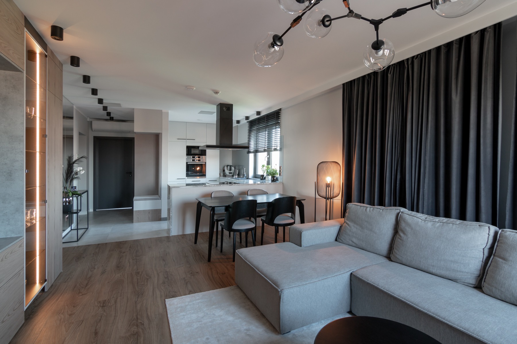 Nowoczesne wnętrze mieszkania z otwartą kuchnią i salonem, szara sofa narożna, drewniana podłoga, czarny stół z krzesłami, oświetlona witryna i designerska lampa podłogowa.