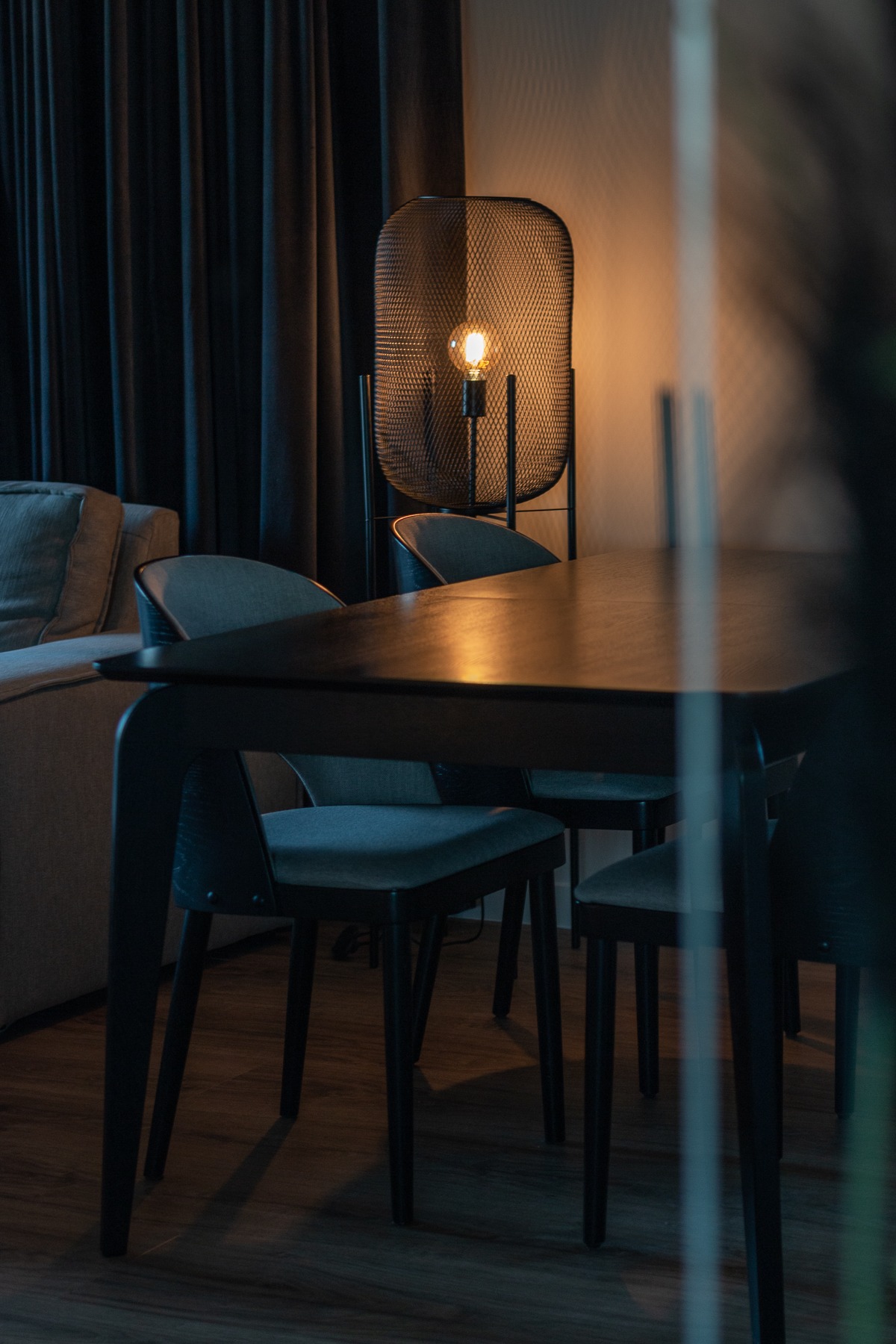 Ciemne, nowoczesne wnętrze jadalni z drewnianym stołem, szarymi krzesłami i lampą z drucianym kloszem, oświetlonym żarówką edisona, na tle zasłoniętego okna.