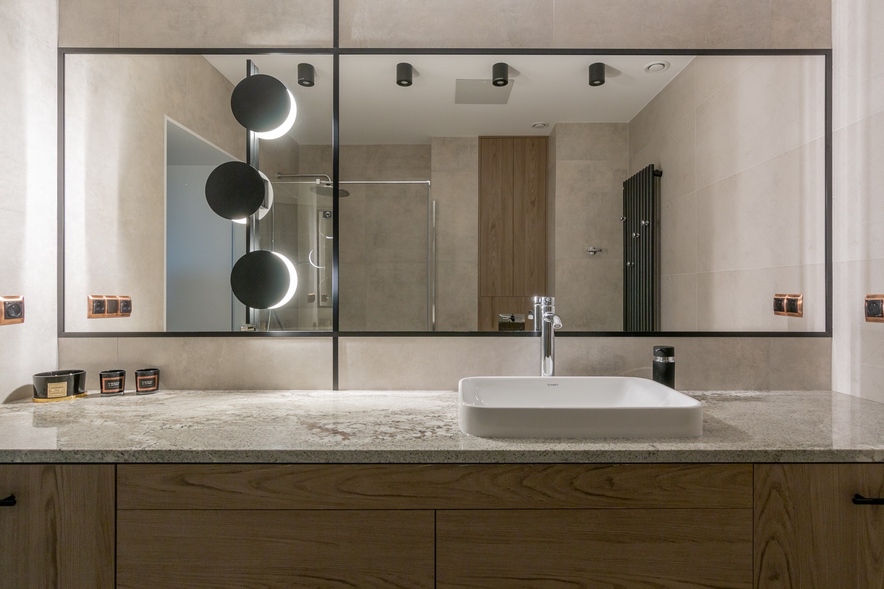 Nowoczesna łazienka z umywalką nablatową, lustrem z czarną ramą i okrągłymi lampami, widoczny prysznic za szybą.