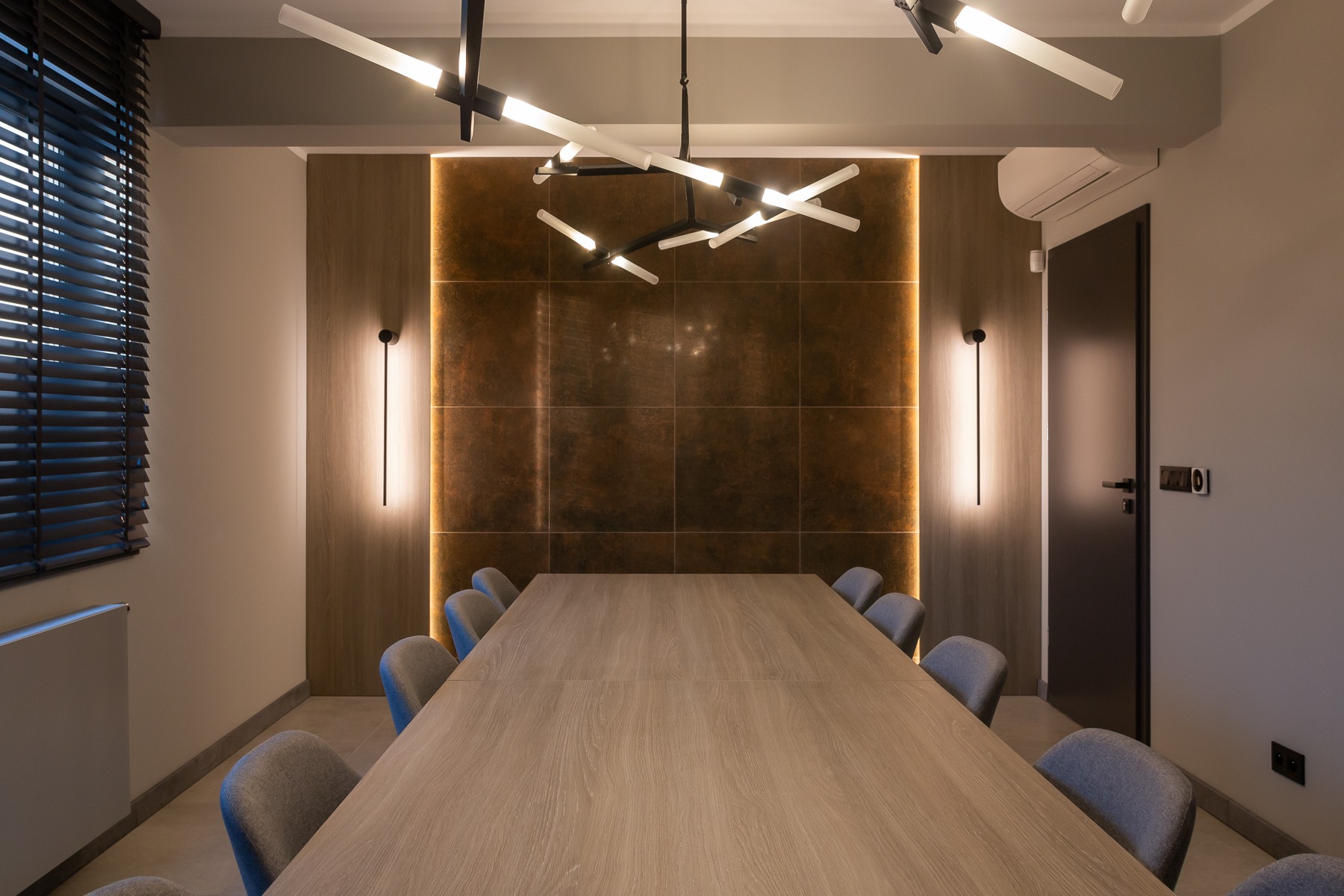 Nowoczesna sala konferencyjna z dużym drewnianym stołem, szarymi krzesłami, designerską lampą sufitową i podświetlaną ścianą z płytek w odcieniach brązu.