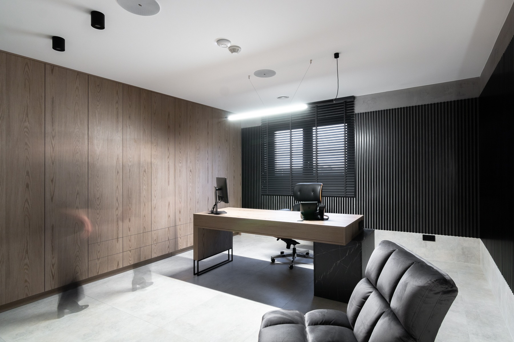 Nowoczesne biuro w Koszalinie z drewnianą ścianą, minimalistycznym biurkiem z marmurowymi elementami, czarnymi żaluzjami i designerskim oświetleniem sufitowym.