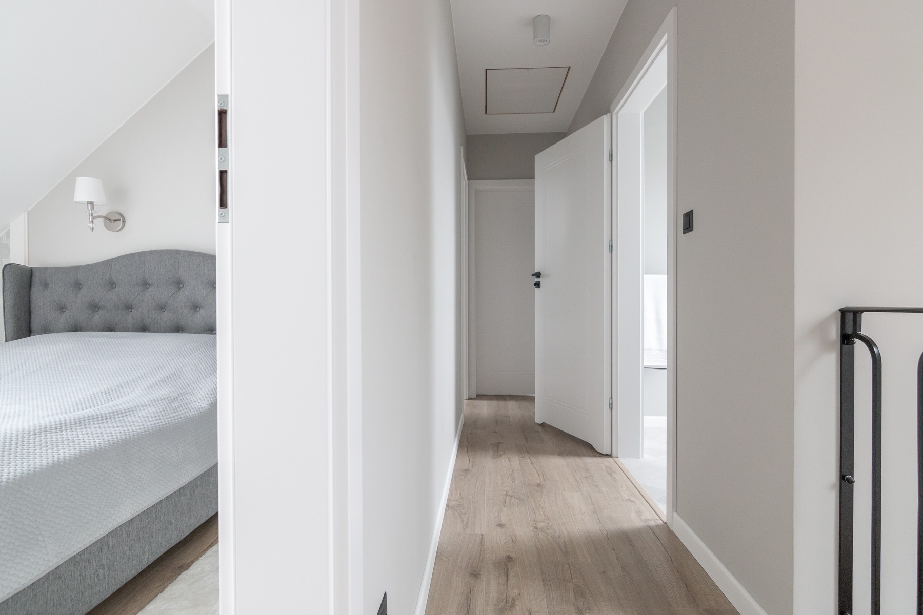 Korytarz z drewnianą podłogą prowadzący do łazienki z otwartymi białymi drzwiami, widoczny fragment sypialni z szarym łóżkiem i lampą ścienną