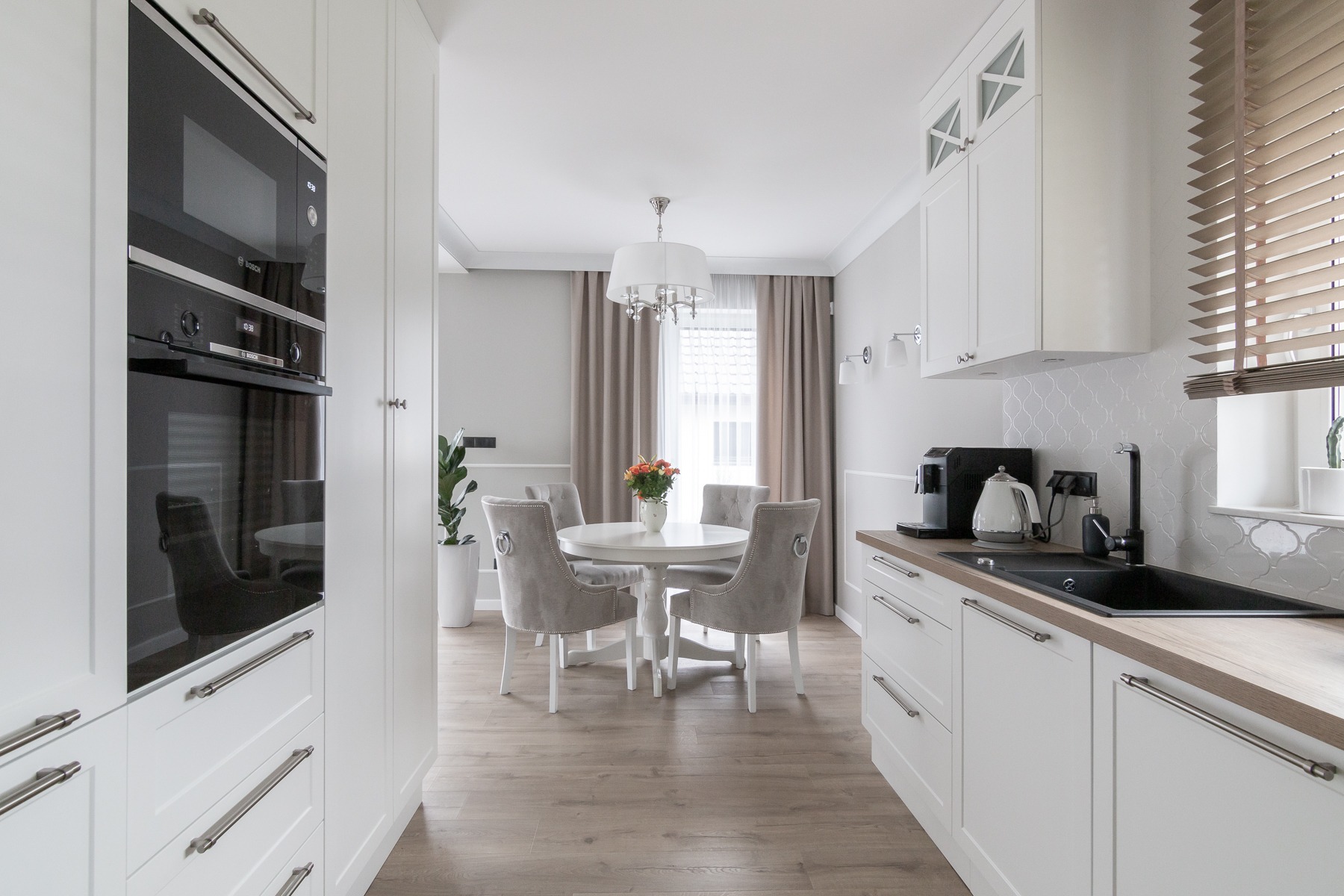Jasna kuchnia z okrągłym stołem, szarymi krzesłami, białymi szafkami i sprzętami AGD marki Bosch.
