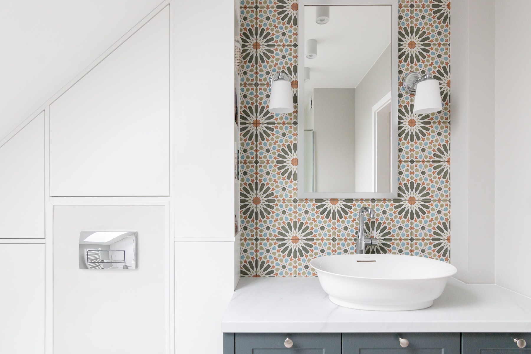 Nowoczesna łazienka z białą, okrągłą umywalką nablatową, baterią chromowaną, lustrem w ramie i ścianą wyłożoną dekoracyjnymi, marokańskimi płytkami w odcieniach beżu, brązu i błękitu. Po lewej...