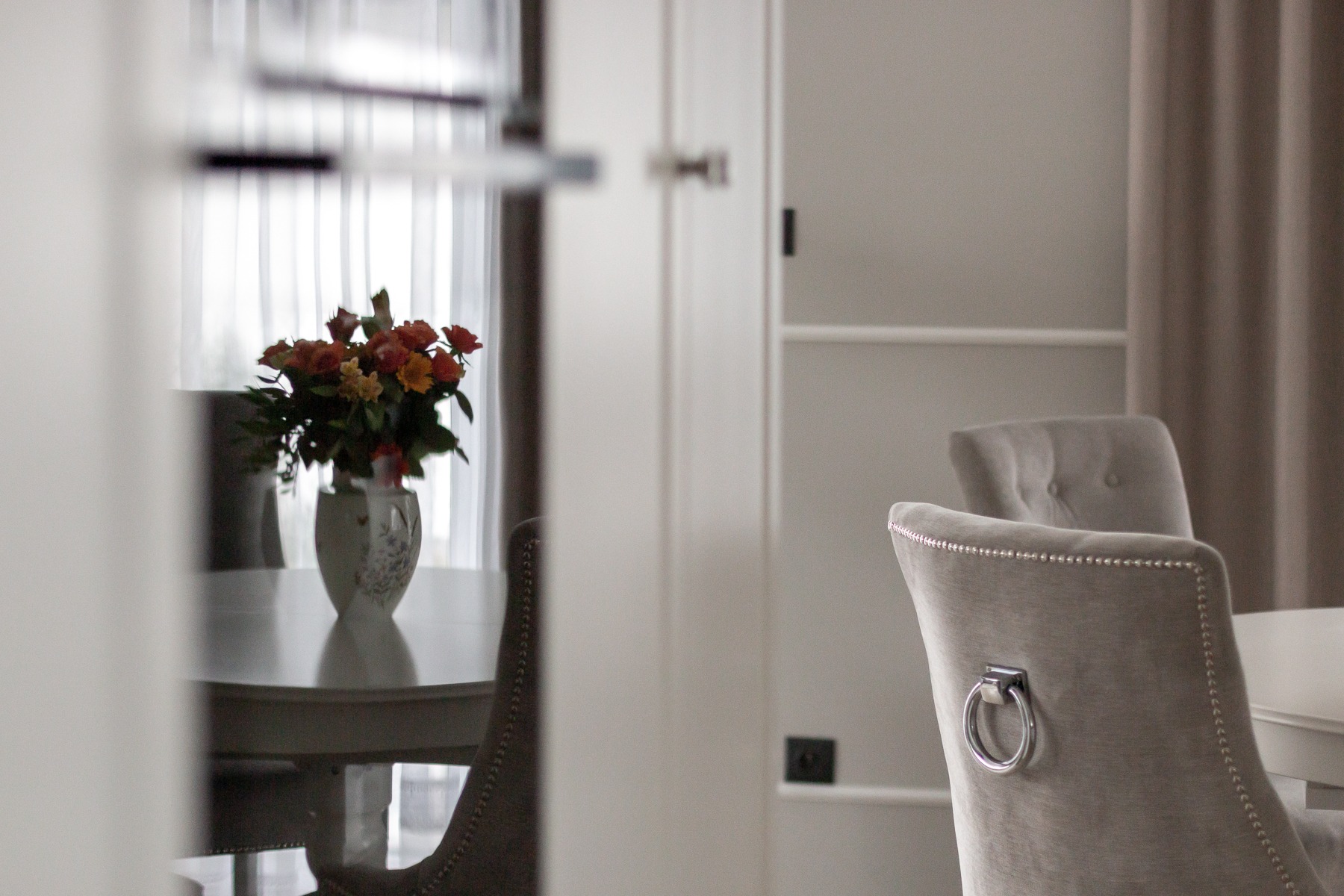 Jasnoszara jadalnia z okrągłym stołem, wazonem z pomarańczowymi różami i krzesłami obitymi aksamitną tkaniną z ozdobnymi gwoździami i metalowymi uchwytami z tyłu.