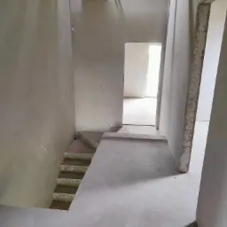 Wnętrze w trakcie prac wykończeniowych: ściany pokryte szarym tynkiem, surowy beton na podłodze i schodach, widoczne kable elektryczne wystające ze ścian, drewniane belki pod sufitem.