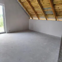 Surowe wnętrze poddasza z drewnianą konstrukcją dachu, ścianami po wstępnym wykończeniu i betonową podłogą, widok na okno.