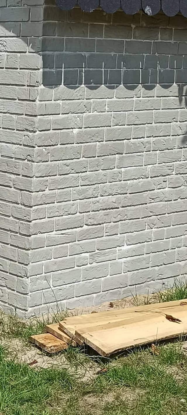 Fragment ściany zewnętrznej budynku wykończonej szarym tynkiem imitującym cegłę, z widocznymi fugami, u podstawy leżą drewniane deski i rośnie trawa.