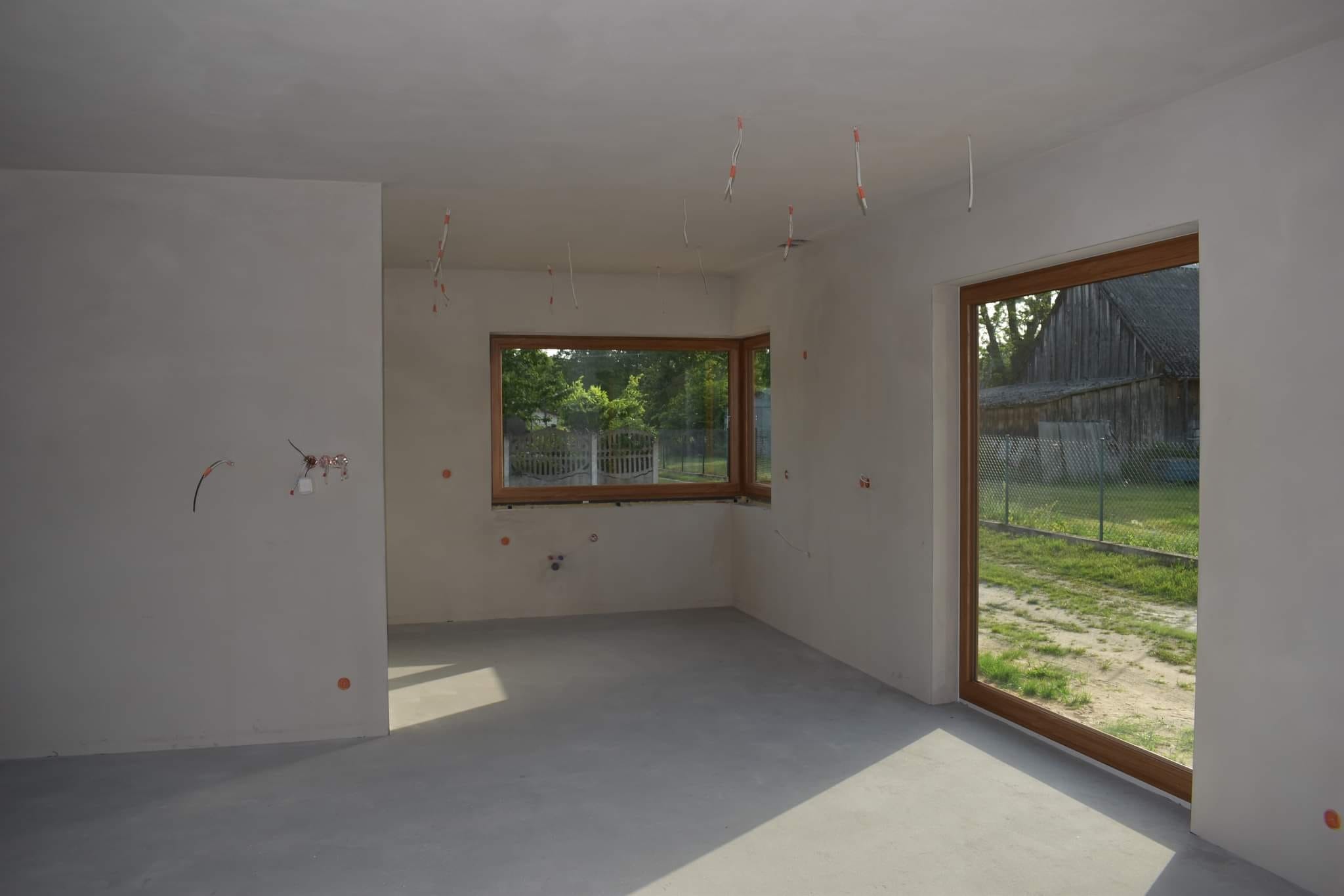 Surowe wnętrze po wstępnych pracach wykończeniowych: gładkie, jasne ściany i wylewka podłogowa, widoczne wyprowadzenia instalacji elektrycznej na ścianach i suficie, okno i drzwi balkonowe...