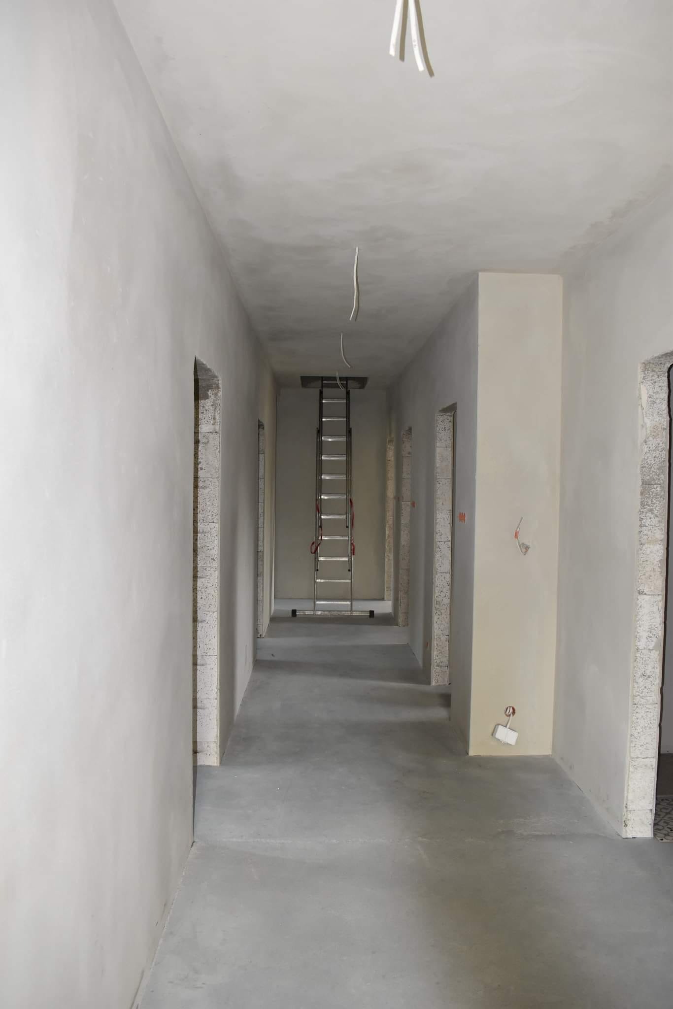 Długi korytarz w trakcie prac wykończeniowych; ściany pokryte świeżym tynkiem, widoczna rozstawiona drabina aluminiowa, wystające przewody elektryczne i niezabezpieczone otwory drzwiowe.