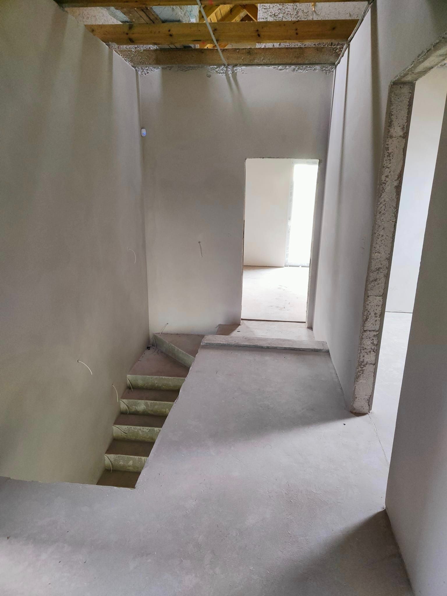 Wnętrze w trakcie prac wykończeniowych: ściany pokryte szarym tynkiem, surowy beton na podłodze i schodach, widoczne kable elektryczne wystające ze ścian, drewniane belki pod sufitem.