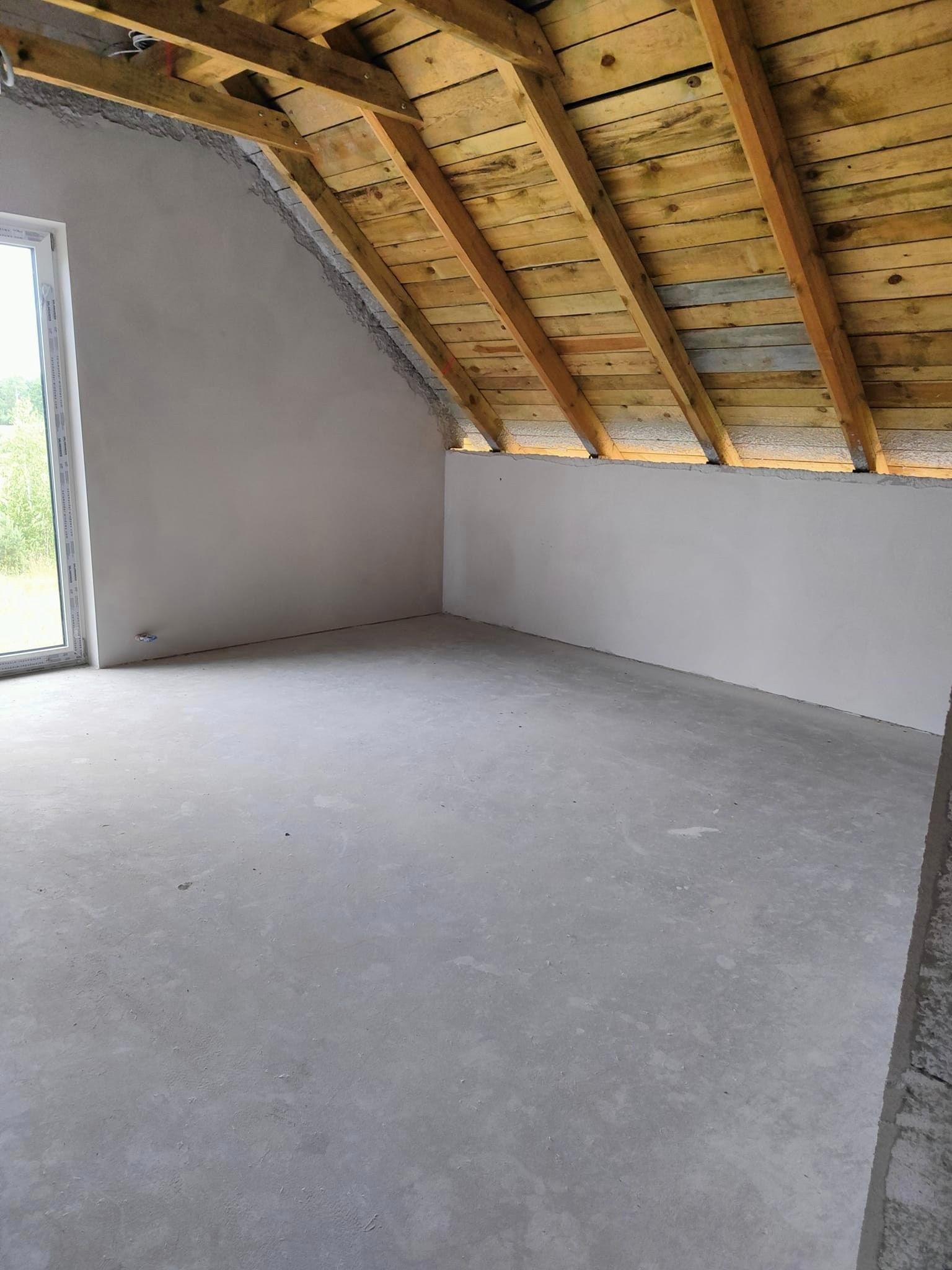 Surowe wnętrze poddasza z drewnianą konstrukcją dachu, ścianami po wstępnym wykończeniu i betonową podłogą, widok na okno.