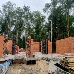 Wznoszenie ścian z czerwonej cegły Porotherm na fundamencie, widoczne szalunki i zbrojenia, w tle las.