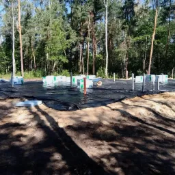 Przygotowany teren pod budowę domu z rozłożoną czarną folią izolacyjną, wystającymi pionowymi rurami i bloczkami fundamentowymi w tle lasu.