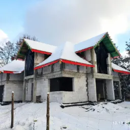 Surowy stan dwukondygnacyjnego domu z białych bloczków w zimowej scenerii, z widocznym deskowaniem dachu w kolorze zielonym i czerwonym, częściowo pokrytym śniegiem.