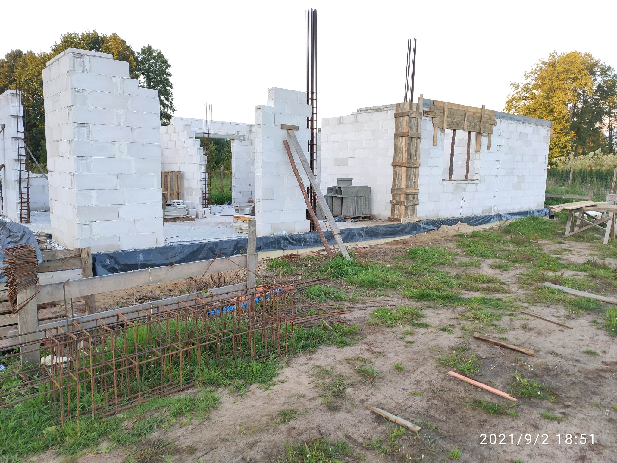 Budowa domu z białych bloczków: ściany w trakcie wznoszenia, widoczne zbrojenia i szalunki okienne, teren budowy z resztkami materiałów i datą wykonania zdjęcia w rogu.