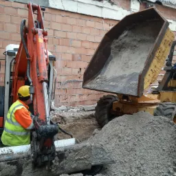 Prace ziemne: operator koparki w kamizelce odblaskowej i kasku, wykop z widoczną rurą, obok wywrotka z ziemią, w tle mur z cegły dziurawki.