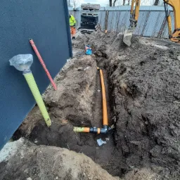 Wykop z widocznymi rurami kanalizacyjnymi z tworzywa sztucznego, częściowo połączonymi, obok ściany budynku i koparki, na placu budowy.