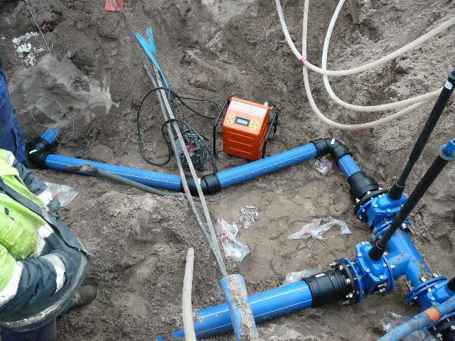 Niebieskie rury wodociągowe z czarnymi złączkami ułożone w wykopie, obok narzędzia i pomarańczowa skrzynka. Widoczne elementy instalacji przyłącza wod-kan.