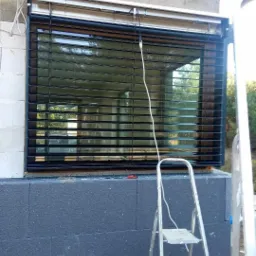 Zamontowane, czarne rolety zewnętrzne na oknie budynku w trakcie prac wykończeniowych elewacji, drabina oparta o ścianę.