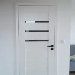 Białe drzwi wewnętrzne z poziomymi, czarnymi przeszkleniami, czarną klamką i widoczną listwą przypodłogową, w minimalistycznym wnętrzu z drewnianymi schodami w tle.