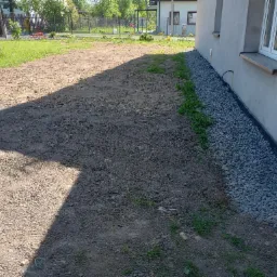 Przygotowany teren pod trawnik z drenażem z kamieni przy ścianie budynku, widoczne okno i rynna.