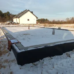 Zimowy widok na fundamenty domu jednorodzinnego z izolacją pionową, na fundamencie leżą plany budowlane przykryte kocem, w tle biały dom i samochód