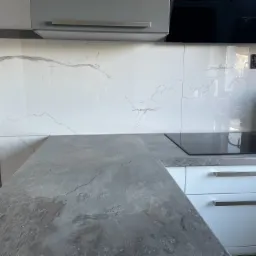 Fragment nowoczesnej kuchni z blatem imitującym kamień łupkowy, białe szafki z poziomymi, metalowymi uchwytami i płytki ścienne z marmurowym wzorem, widoczna płyta indukcyjna.