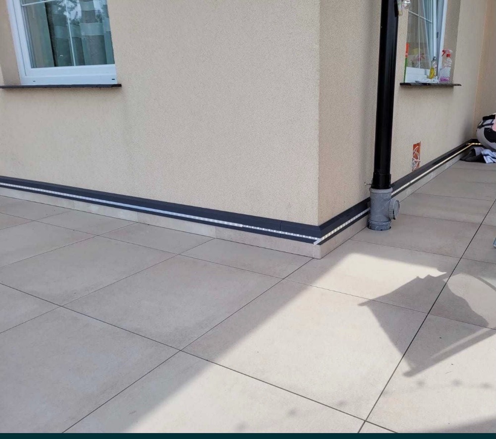 Elewacja domu z zamontowanym oświetleniem LED wzdłuż cokołu, widoczny róg budynku z rurą spustową i fragment okna.