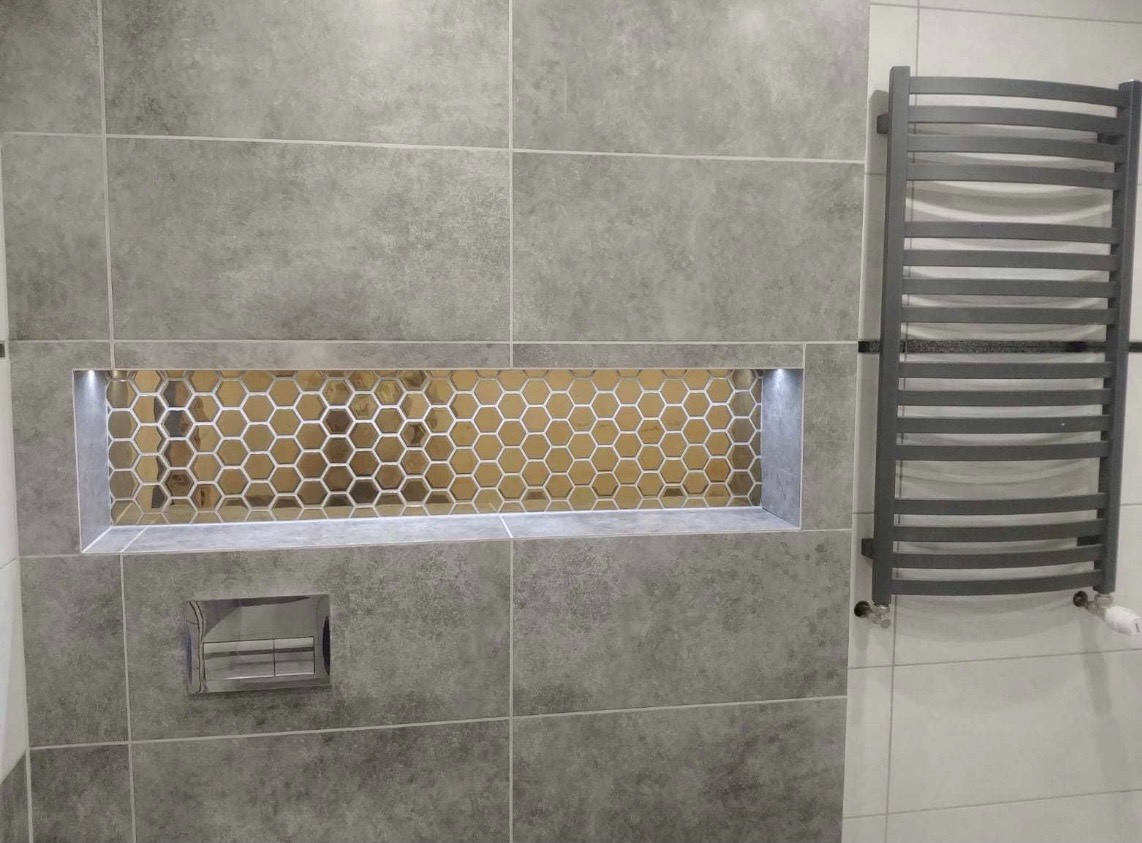 Szare płytki w łazience z wnęką wyłożoną mozaiką w kształcie plastra miodu, podświetloną od góry, obok grzejnik drabinkowy.