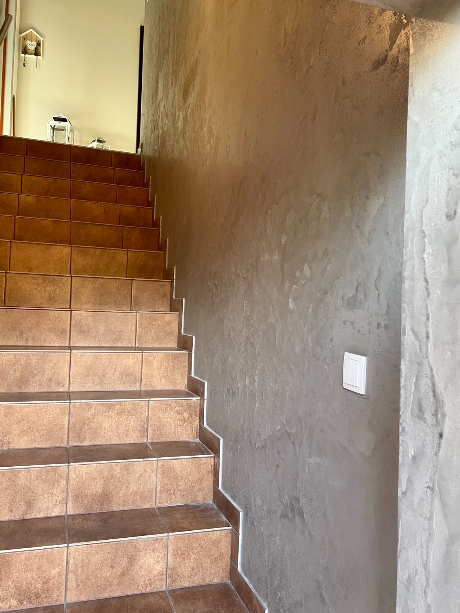 Klatka schodowa z brązowymi płytkami, ściana wykończona betonem architektonicznym w odcieniach szarości, widoczny włącznik światła.