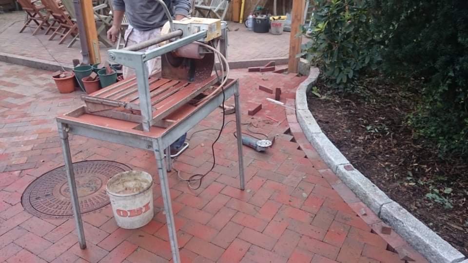 Cięcie cegieł klinkierowych na zewnątrz budynku za pomocą przecinarki stołowej. Widoczne fragmenty ułożonej nawierzchni z cegły, obrzeże trawnika z kamienia, szlifierka kątowa oraz wiadro.