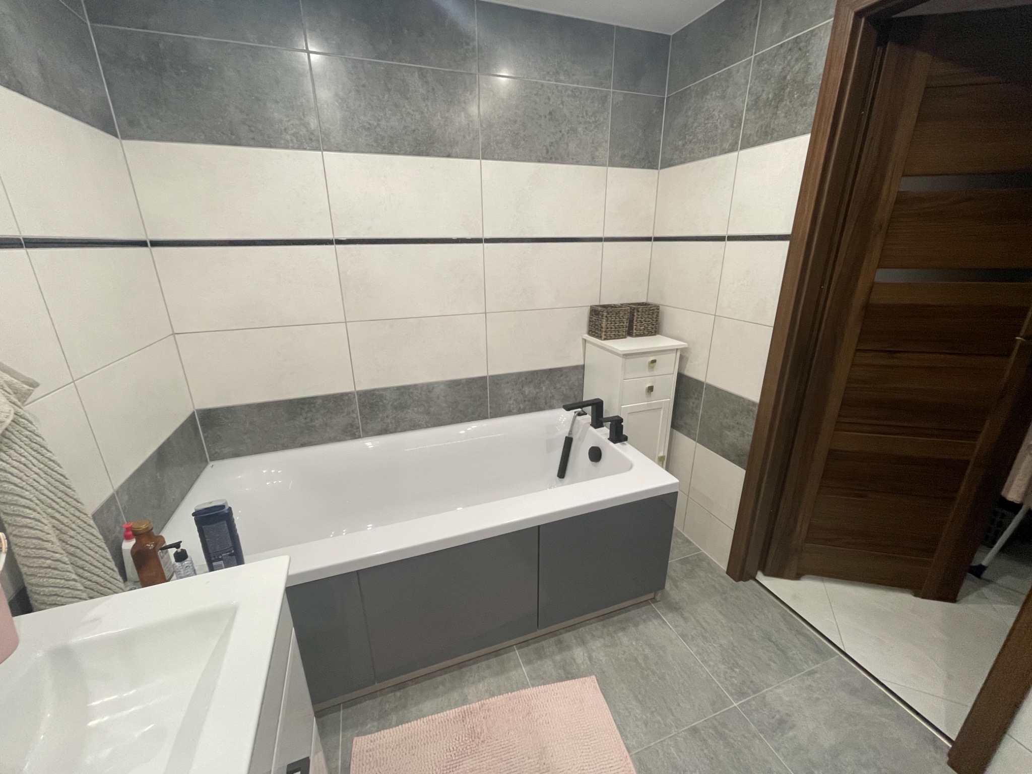 Nowoczesna łazienka z wanną obudowaną szarą zabudową, białą ceramiką, szaro-białymi płytkami i drewnianymi drzwiami, widok na fragment umywalki i dywanik.