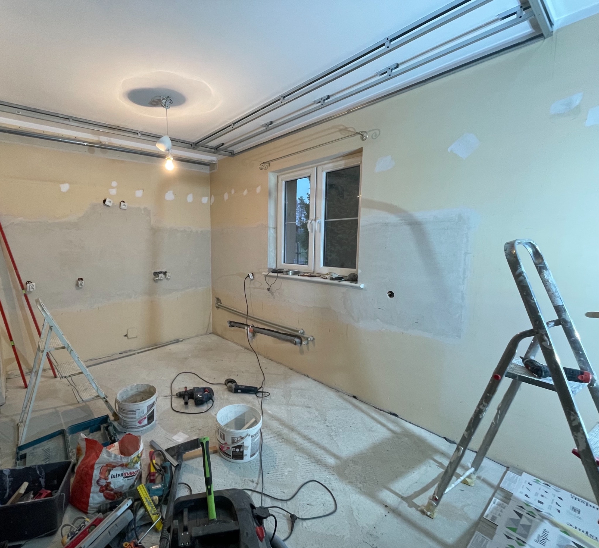 Wnętrze pokoju w trakcie prac wykończeniowych: ściany z częściowo nałożoną gładzią, widoczne instalacje elektryczne, okno, drabiny i narzędzia malarskie na podłodze.